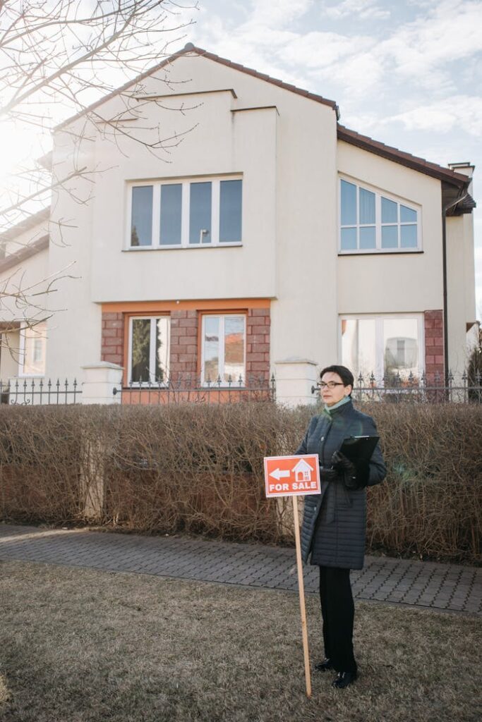 Real estate agent stands with a 'For Sale' sign outside a house on a sunny day, ready for potential buyers.